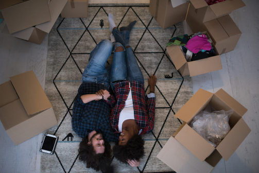 young interacial couple lay on their backs smiling surrounded by moving boxes after their relocation to nevada